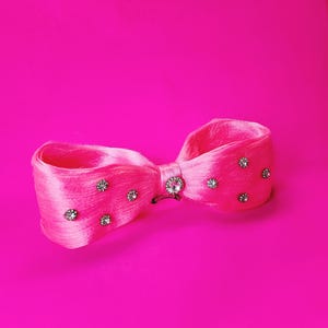 May include: A bright pink hair bow with a satin finish, adorned with sparkling rhinestones. The bow features a central rhinestone embellishment and smaller rhinestones scattered across the loops. The background is a solid, vibrant pink.