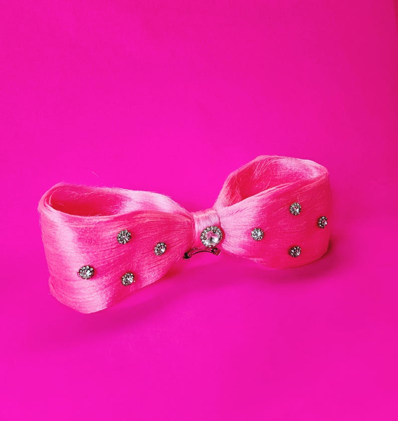 May include: A bright pink hair bow with a satin finish, adorned with sparkling rhinestones. The bow features a central rhinestone embellishment and smaller rhinestones scattered across the loops. The background is a solid, vibrant pink.