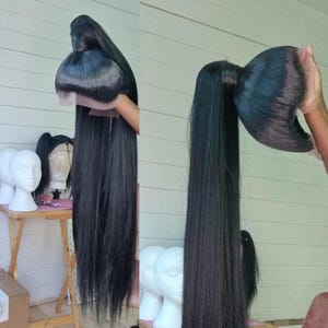 May include: A long, black wig with a full crown and long, straight hair. The wig is held up, displaying its length and style. Several mannequin heads are in the background, indicating a wig shop or styling setup.