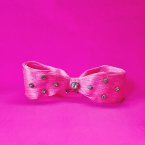 May include: A bright pink hair bow with a bow tie design. The bow is adorned with small, sparkling rhinestones and a larger, central rhinestone. The background is a solid, vibrant pink.