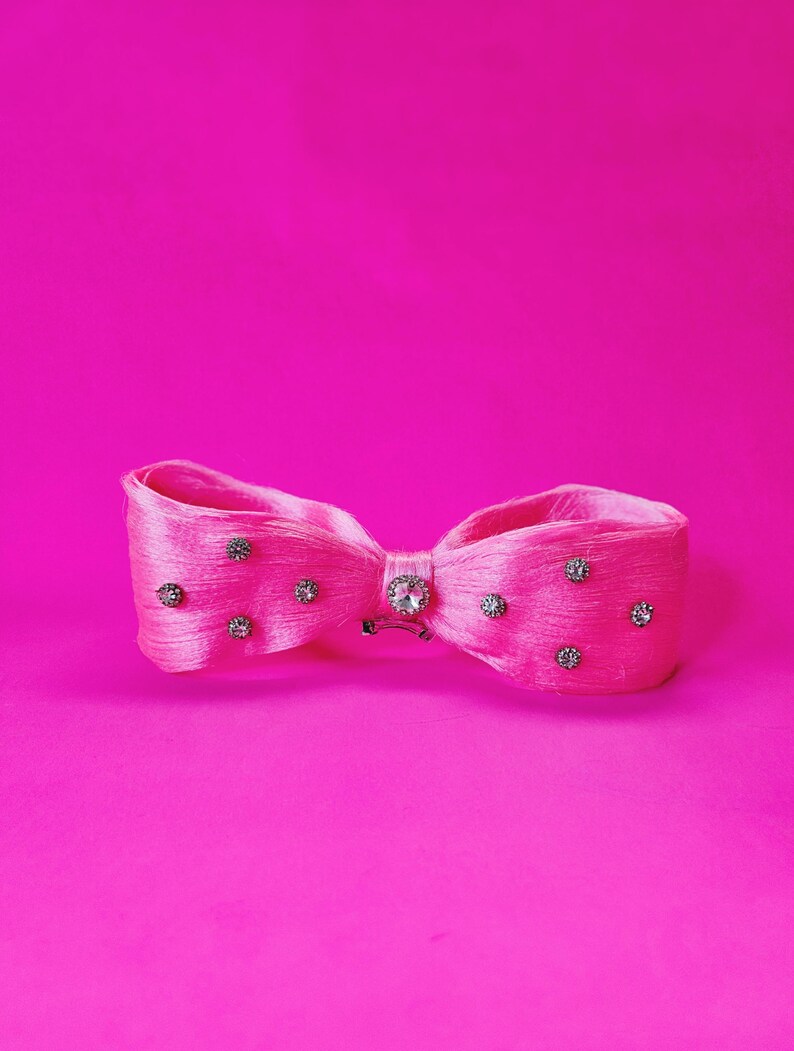 May include: A bright pink hair bow with a bow tie design. The bow is adorned with small, sparkling rhinestones and a larger, central rhinestone. The background is a solid, vibrant pink.
