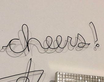 May include: A black wire sculpture of a martini glass and the word "cheers" on a white wall. A silver framed mirror with a tiled border is to the right of the sculpture.