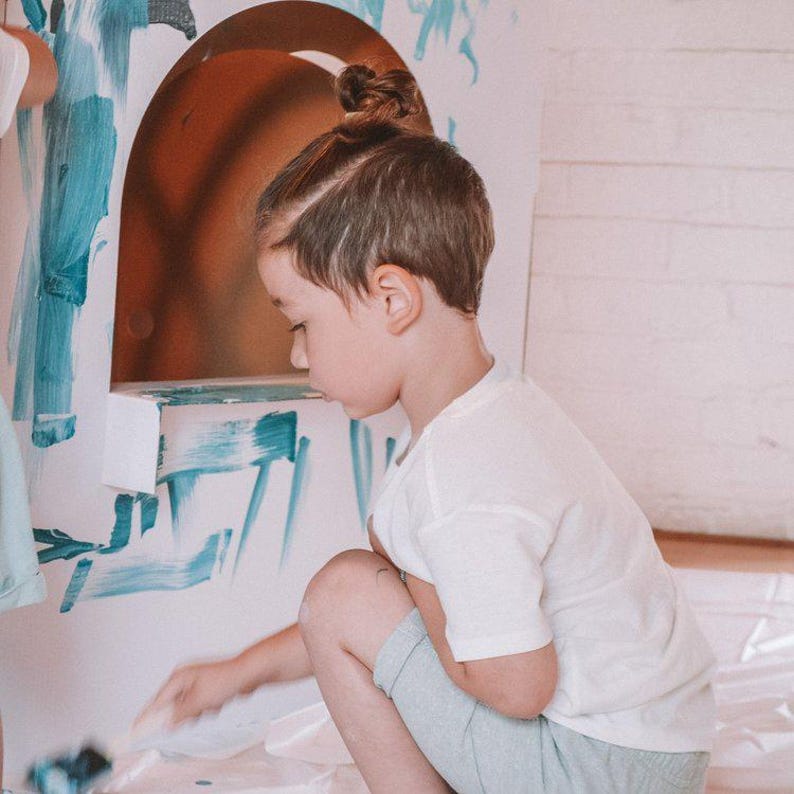 K&ouml;nnte beinhalten: Ein kleines Kind mit braunen Haaren in einem Dutt malt an einer wei&szlig;en Wand mit blauer Farbe. Das Kind tr&auml;gt ein wei&szlig;es T-Shirt und eine hellblaue Shorts.