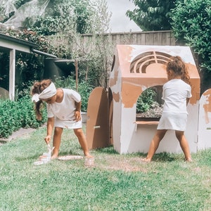 K&ouml;nnte beinhalten: Zwei Kinder bemalen ein Spielhaus aus Pappe in einem Garten. Das Spielhaus ist wei&szlig; mit braunen Akzenten und hat eine T&uuml;r und ein Fenster. Die Kinder tragen wei&szlig;e Hemden und Shorts. Das Gras ist gr&uuml;n.