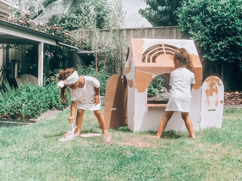 K&ouml;nnte beinhalten: Zwei Kinder bemalen ein Spielhaus aus Pappe in einem Garten. Das Spielhaus ist wei&szlig; mit braunen Akzenten und hat eine T&uuml;r und ein Fenster. Die Kinder tragen wei&szlig;e Hemden und Shorts. Das Gras ist gr&uuml;n.