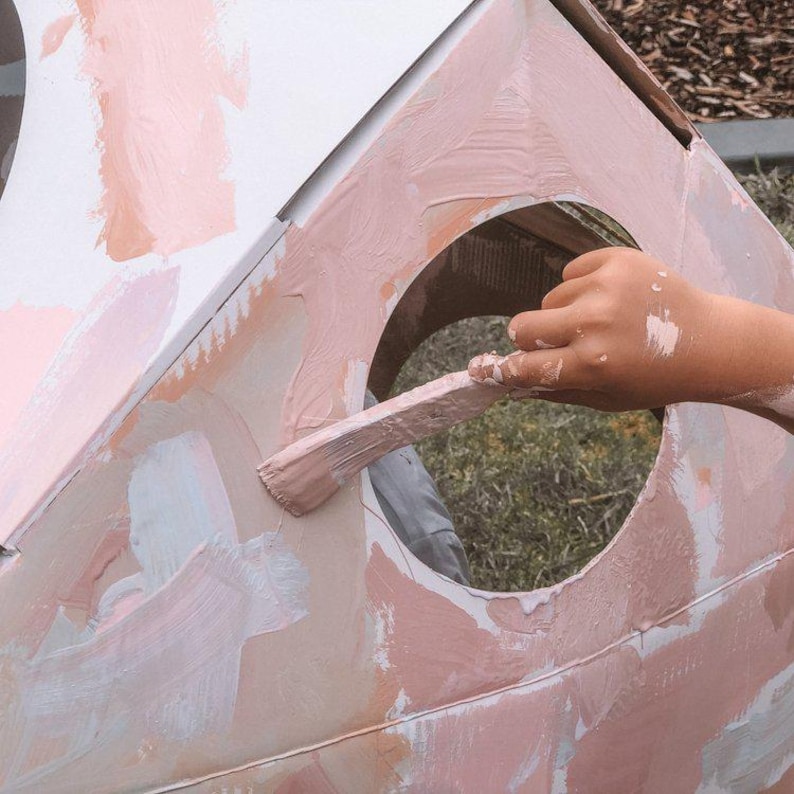 K&ouml;nnte beinhalten: Eine Person malt ein rosa Karton-Spielhaus mit einem wei&szlig;en Pinsel. Das Spielhaus hat eine runde Fenster&ouml;ffnung.
