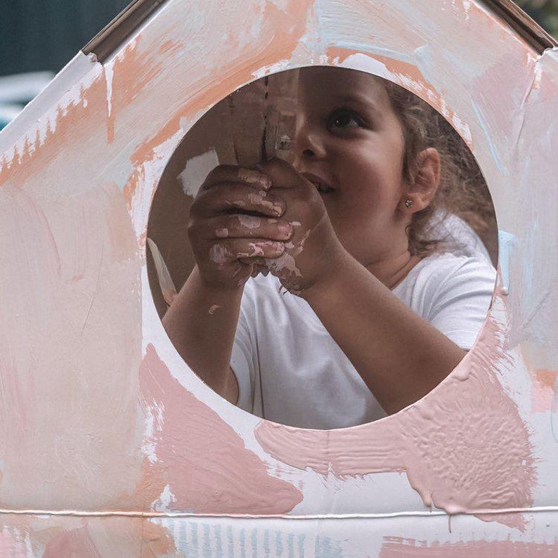 K&ouml;nnte beinhalten: Ein junges Kind mit Farbe an den H&auml;nden schaut aus einem runden Fenster in einem Kartonhaus, das in Rosa-, Wei&szlig;- und Blaut&ouml;nen gestrichen ist.