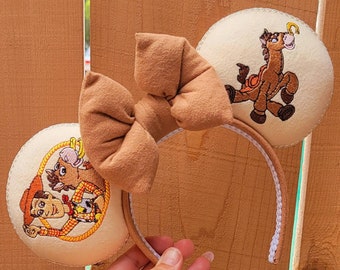 Può includere: Una fascia per capelli color cuoio con orecchie ricamate con personaggi di Toy Story e un grande fiocco. Le orecchie presentano immagini di Woody, Bullseye e altri personaggi del film. Il fiocco è realizzato in un tessuto morbido e strutturato.