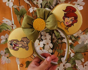 May include: Yellow fabric headband with two circular ears featuring embroidered character designs. The left ear shows a brown puppy, the right ear shows a woman with red hair, and a central olive green fabric bow features a yellow star-shaped badge with the text Together In Paris.