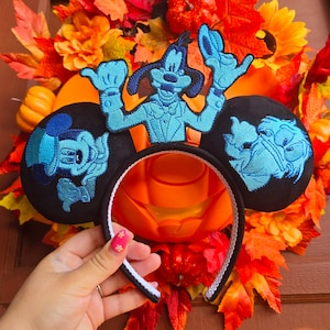 May include: Black mouse ear headband featuring embroidered cartoon characters in shades of blue. The headband is set against a backdrop of fall foliage, pumpkins, and flowers, creating a festive autumn theme.