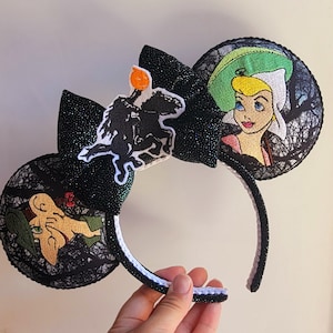 May include: A black headband featuring two circular ears covered in black lace. The left ear displays an embroidered character with a green hat, while the right ear shows a character in a green bonnet. A black glittery bow sits in the center with an embroidered headless horseman figure.