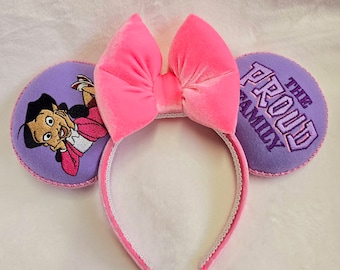 Può includere: Una fascia per capelli con fiocco in velluto rosa e dettagli orecchie viola. Un orecchio presenta un'immagine ricamata di un personaggio dei cartoni animati, l'altro orecchio mostra la scritta "THE PROUD FAMILY" in ricamo viola. La fascia è rifinita con pizzo bianco.