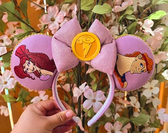 May include: A lavender headband with embroidered character ears. The ears feature Ariel and Hercules designs. A glittery pink bow and a gold emblem with a lightning bolt complete the design. The headband is held by a hand.