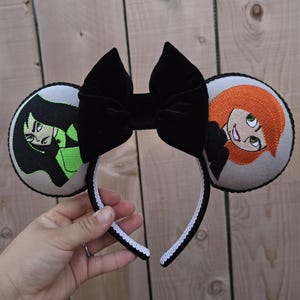 May include: A black headband with two round gray ears featuring embroidered cartoon characters. A large black bow sits atop the headband. The headband is trimmed with white lace.