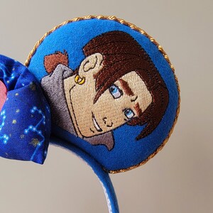 May include: Blue headband with embroidered character art. The headband features a blue bow with a constellation pattern and a pink heart. The character art is a circular patch with a gold trim, depicting a cartoon character with brown hair and blue eyes.