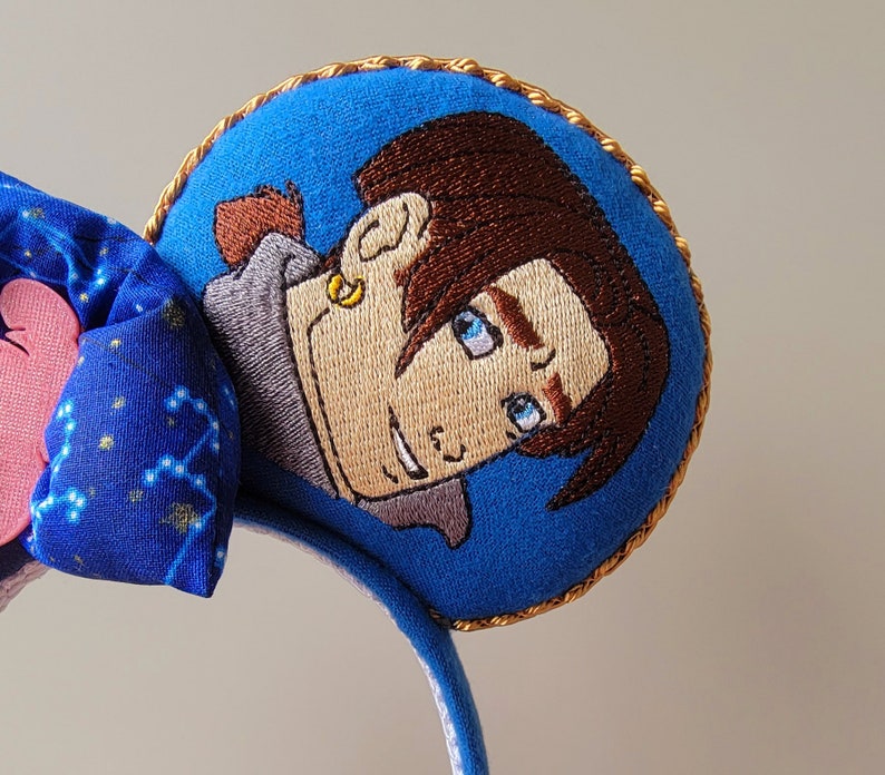 May include: Blue headband with embroidered character art. The headband features a blue bow with a constellation pattern and a pink heart. The character art is a circular patch with a gold trim, depicting a cartoon character with brown hair and blue eyes.