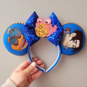 May include: Embroidered mouse ears with a blue bow patterned with constellations. The ears are blue with gold trim and feature embroidered characters. A pink and yellow character is on the bow.