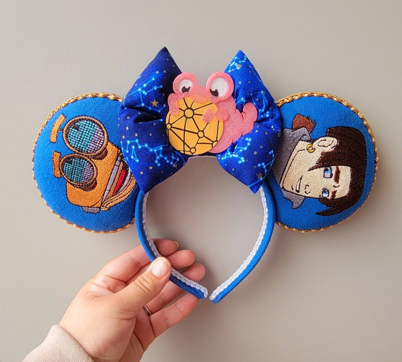 May include: Embroidered mouse ears with a blue bow patterned with constellations. The ears are blue with gold trim and feature embroidered characters. A pink and yellow character is on the bow.