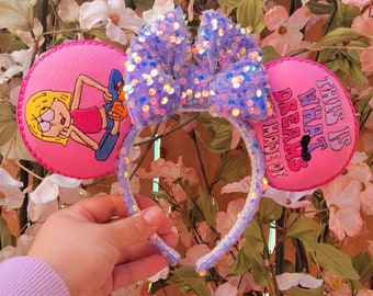 Può includere: Una fascia per capelli di lavanda con paillettes con un grande fiocco e due orecchie circolari rosa. Un orecchio presenta un personaggio dei cartoni animati, l'altro recita "THIS IS WHAT DREAMS ARE MADE OF". La fascia è contro uno sfondo floreale.