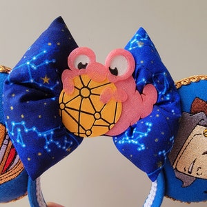 May include: A blue fabric headband with a constellation pattern, featuring a pink crab holding a yellow sphere. The headband has a white trim and gold accents, with embroidered details on the sides.