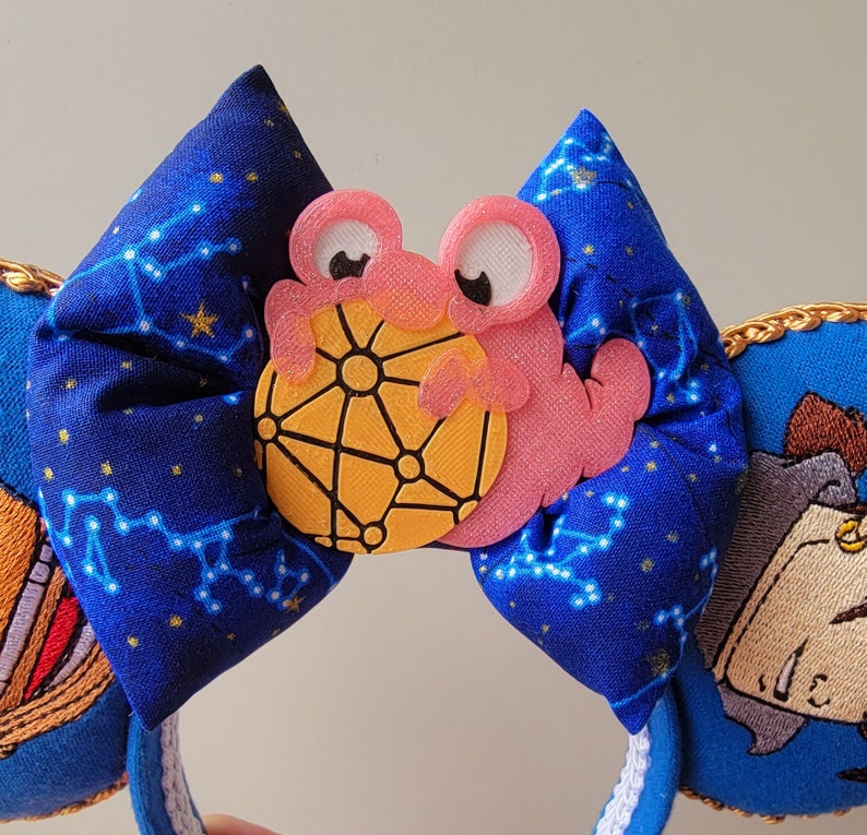 May include: A blue fabric headband with a constellation pattern, featuring a pink crab holding a yellow sphere. The headband has a white trim and gold accents, with embroidered details on the sides.