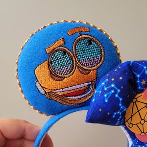 May include: Blue character-themed mouse ears with a gold trim. The character's face is embroidered in orange and brown, with mesh-like eyes. A blue bow with a constellation pattern and a pink and yellow accent complete the design. The headband is blue.