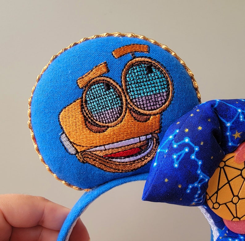 May include: Blue character-themed mouse ears with a gold trim. The character's face is embroidered in orange and brown, with mesh-like eyes. A blue bow with a constellation pattern and a pink and yellow accent complete the design. The headband is blue.