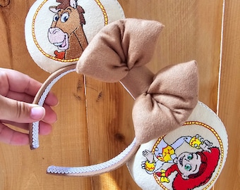 Può includere: Un paio di orecchie da topo con immagini ricamate di un cavallo dei cartoni animati e una cowgirl. Le orecchie sono color crema con un fiocco marrone e un bordo in pizzo bianco sulla fascia. Il design è ispirato a un popolare film d'animazione.