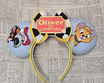 May include: A headband featuring two light blue circular ears with embroidered character designs. The left ear shows a dog, the right ear shows a kitten, and a central yellow and black bow displays a red plaque with the text Oliver AND COMPANY.