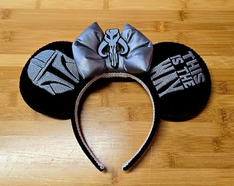 May include: Black mouse ears with silver embroidered designs and a silver bow. One ear features a Mandalorian helmet design, the other reads "This is the Way." The headband is black with a white trim.