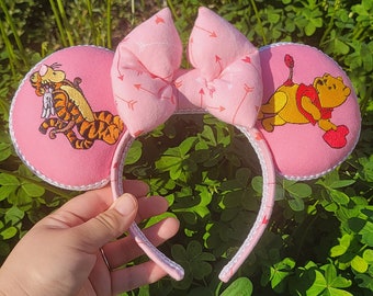 May include: Pink mouse ear headband with Tigger and Winnie the Pooh designs. The ears have white trim and the bow is light pink with arrow patterns. A fun accessory for fans of the classic characters.