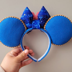 May include: Blue mouse ear headband with gold trim and a bow featuring a constellation pattern. The bow has a pink accent in the centre. The ears are a solid blue colour.