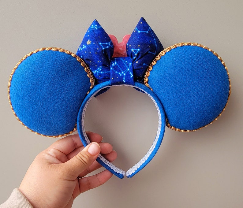May include: Blue mouse ear headband with gold trim and a bow featuring a constellation pattern. The bow has a pink accent in the centre. The ears are a solid blue colour.