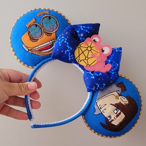 May include: Character-themed ear headband in blue with embroidered details. The ears feature cartoon characters and are trimmed with gold. A blue bow with a constellation pattern and a pink character accent the headband.