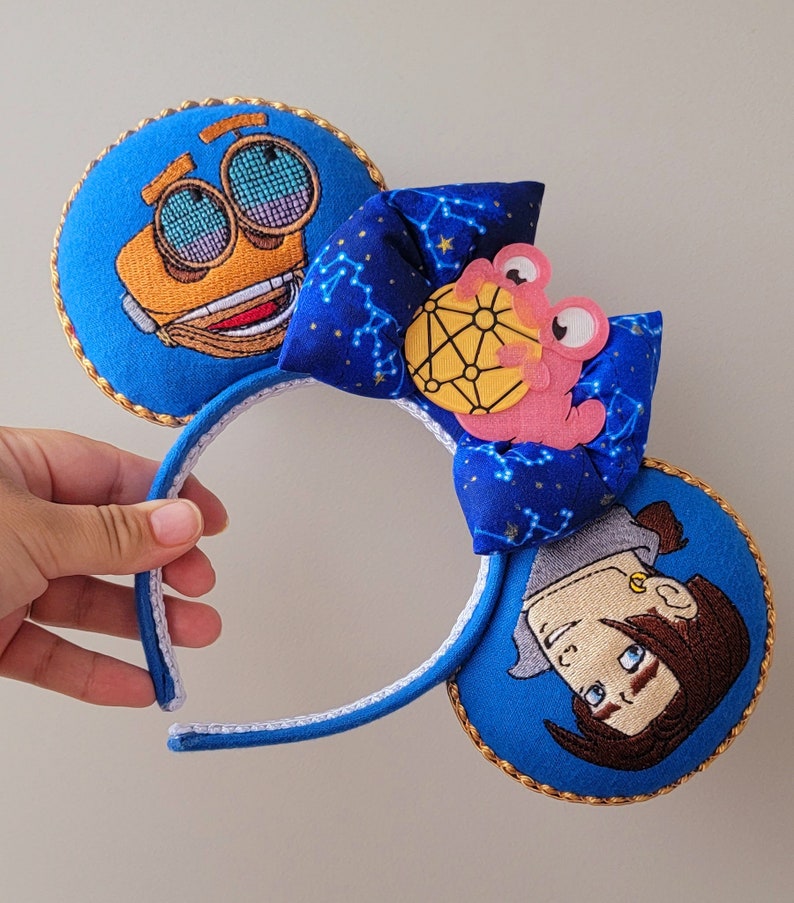 May include: Character-themed ear headband in blue with embroidered details. The ears feature cartoon characters and are trimmed with gold. A blue bow with a constellation pattern and a pink character accent the headband.