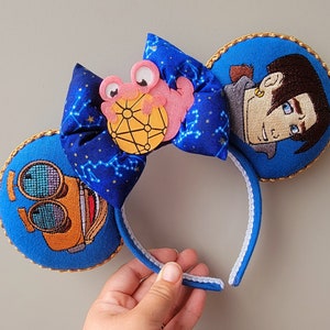 May include: Blue headband with embroidered character ears. One ear features a character's face, the other a machine. A blue bow with a constellation pattern and a pink character is centered on the headband. Gold trim outlines the ears.