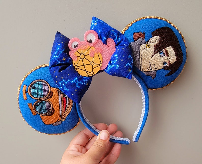 May include: Blue headband with embroidered character ears. One ear features a character's face, the other a machine. A blue bow with a constellation pattern and a pink character is centered on the headband. Gold trim outlines the ears.