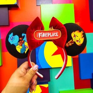 May include: A red velvet bow headband with the text "The Fireplace" on a brown sign. The headband features black circular ear accents with embroidered cartoon characters. The headband is held up against a colorful background.