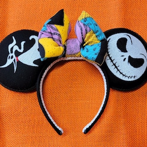 May include: Black mouse ears headband featuring Zero, Jack Skellington, and Sally's patchwork bow. The ears are black with white embroidered designs. The bow is a patchwork of yellow, purple, and blue fabrics. The headband is black with a white trim.