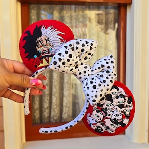May include: A pair of mouse ears featuring a white bow with black spots and red ear accents. One ear displays an embroidered image of Cruella de Vil, while the other shows a group of dalmatian puppies. The headband is white with black spots.