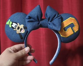 May include: Denim-style mouse ears with a large blue bow. One ear features an embroidered image of Goofy, the other has a gold letter "G". The headband is blue with white stitching.