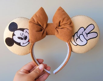 Può includere: Una fascia beige con orecchie da topo marroni e un fiocco marrone. Un orecchio presenta Mickey Mouse, l'altro un segno di pace. La fascia è rifinita con pizzo bianco.