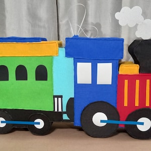 May include: A colorful felt train pinata with a green boxcar, blue engine, and red caboose. The pinata has black wheels, yellow accents, and a white smoke plume.