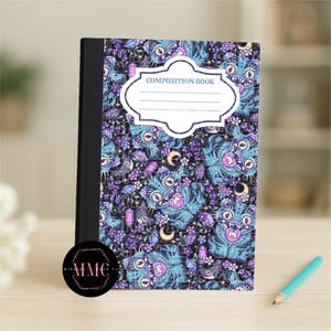 May include: Composition book with a black spine and a colorful cover featuring a repeating pattern of blue and purple fantasy creatures, crystals, and moons. The cover has a white label that says "COMPOSITION BOOK". A blue pencil rests on the table.