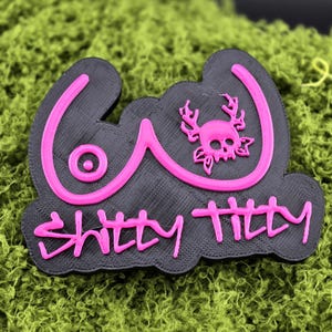 May include: A black and pink 3D-printed patch with the words "SHITTY TITTIES" in pink, along with a stylized breast design and a skull with antlers. The patch is set against a green, textured background.