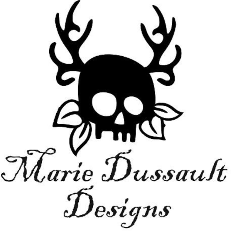 May include: A black graphic decal featuring a skull with large antlers and leaves on either side. Below the graphic, the text Marie Dussault Designs is written in a black serif font. The entire design is presented on a plain white background.