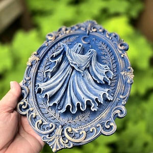May include: An oval-shaped wall plaque featuring a relief sculpture of two ghosts draped in flowing fabric. The piece is finished in a deep blue with gold-toned highlights on the ornate, scrollwork frame and the central crescent moon motif.