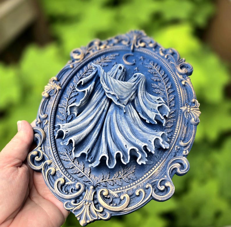 May include: An oval-shaped wall plaque featuring a relief sculpture of two ghosts draped in flowing fabric. The piece is finished in a deep blue with gold-toned highlights on the ornate, scrollwork frame and the central crescent moon motif.