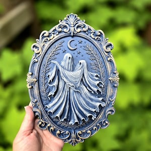 May include: An oval wall plaque featuring a relief sculpture of two ghosts dancing under a crescent moon and stars. The piece is finished in a dark blue and gold color scheme with an ornate, decorative frame surrounding the central scene.