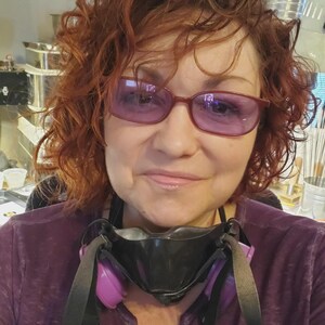 May include: A person with curly auburn hair wearing purple-tinted glasses and a black respirator mask with purple filter cartridges. The individual is dressed in a purple long-sleeved shirt, with the mask resting around the neck area.